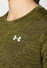 Primer plano de una persona que lleva una camiseta Under Armour texturizada de color verde oliva con un logo blanco en el pecho, mostrando el cuello y la parte superior del torso.