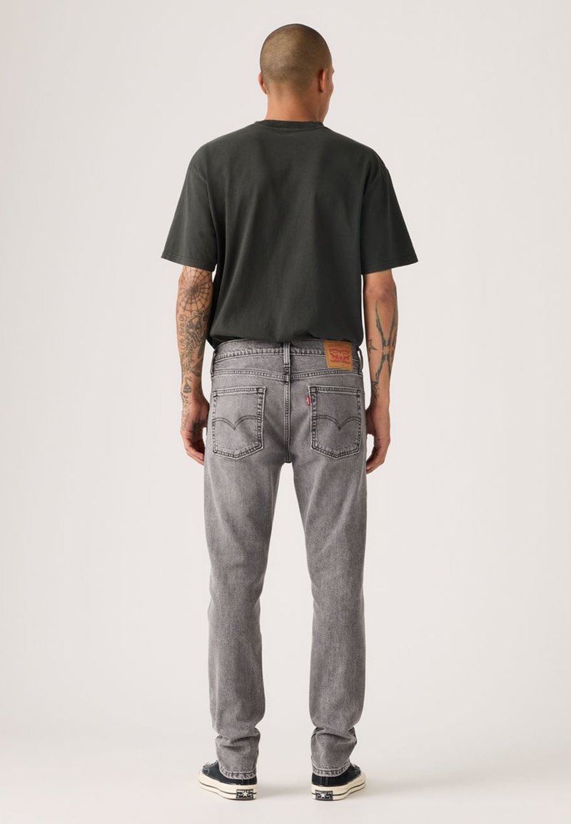 Vaqueros de mezclilla gris con corte de pierna recta, que cuentan con bolsillos traseros y una etiqueta de cuero marrón en la cintura. Combinados con una camiseta negra y zapatillas deportivas.