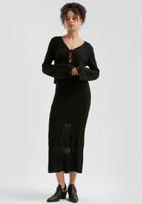 Schwarzes, gestricktes Zweiteiler-Set bestehend aus einer verkürzten Strickjacke mit Knotenverschluss und langen Ärmeln, kombiniert mit einem taillierten, knielangen Rock.