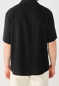 Camicia nera a maniche corte in tessuto testurizzato, con taglio dritto e colletto standard. Presenta un giromanica sul retro e spacchi laterali all'orlo.