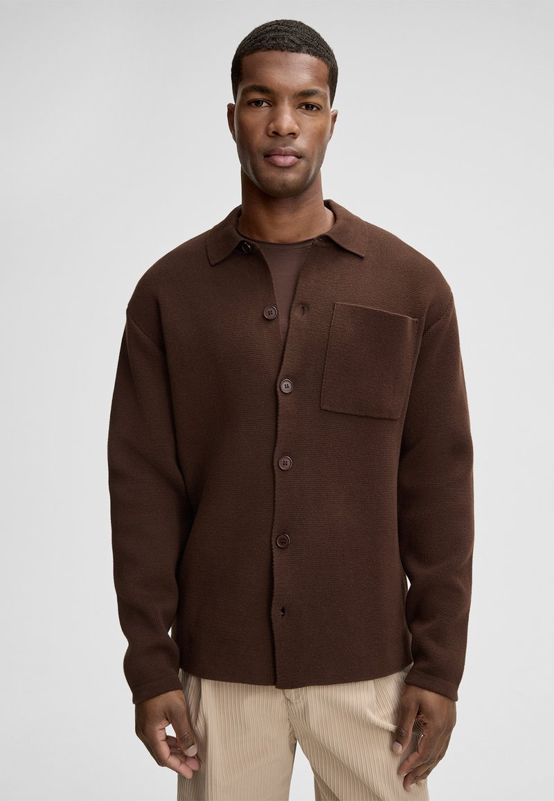 Brązowa dzianinowa koszula zapinana na guziki z kołnierzykiem, przednią kieszonką i ciemnymi guzikami; noszona na ciemnym topie i zestawiona z jasnymi wyrównanymi spodniami.