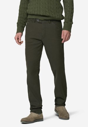 Mann trägt dunkelgrüne Hose, olivgrünen Zopfpullover und hellbraune Wildlederschuhe, steht auf weißem Hintergrund.