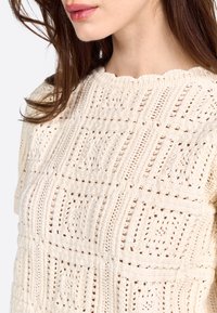 Suéter de color crema tejido, con patrones texturizados, que presenta secciones de tejido abierto, cuello de canalé y borde ondulado. Ajuste casual.
