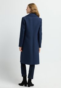 Cappotto blu navy con un motivo trapuntato e testurizzato, caratterizzato da una silhouette lunga, colletto a scollatura e maniche sartoriali, abbinato a jeans scuri e stivaletti alla caviglia.