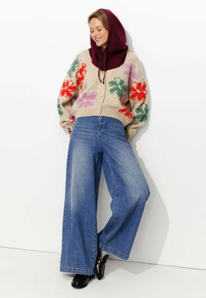 Cardigan en tricot beige avec des motifs floraux rouges, verts et violets ; associé à un jean bleu à jambes larges et des chaussures noires. Accessoire à capuche bordeaux.
