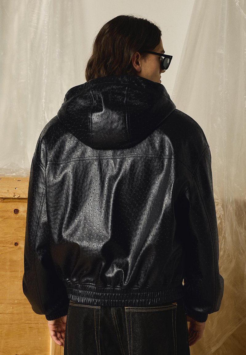 Person wearing black textured leather jacket with hood, dark sunglasses, and dark pants, standing with back facing camera indoors.