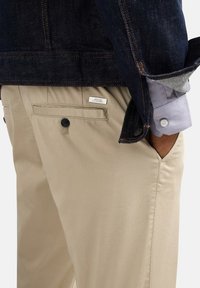 Pantaloni beige con finitura testurizzata, dotati di tasche posteriori con bottoni neri e un piccolo tag con il marchio. Giacca in denim scuro e camicia grigia visibili.