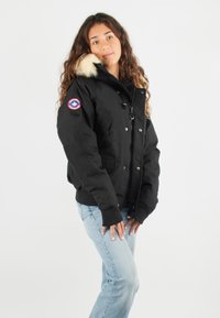 Veste isolée noire avec une capuche doublée de fourrure, fermeture à pression à l'avant, deux poches latérales et un patch logo sur la manche gauche.