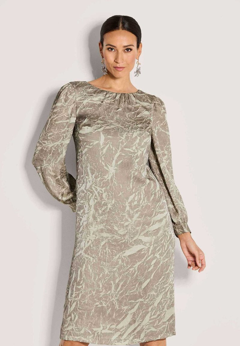 Frau in einem langärmligen, knielangen Kleid mit einem beigen und hellgrünen abstrakten Muster, die vor einem neutralen Hintergrund steht.