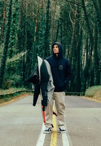 Sweat à capuche bleu marine avec un logo, pantalon beige et baskets noires et blanches. Tenant des vestes grises et noires sur une route forestière.