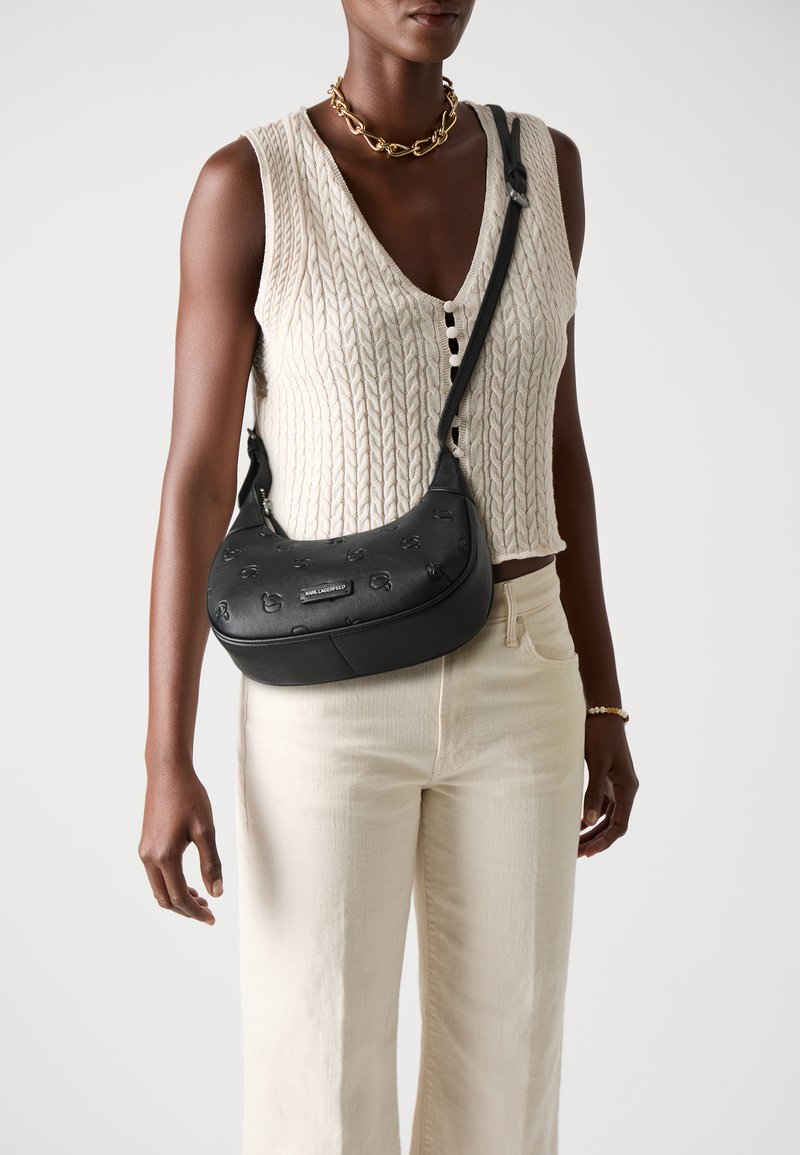 Borsa nera con motivo stampato, forma curvata e chiusura con zip. Indossata a tracolla con una lunga cinghia, abbinata a un top in maglia color crema e pantaloni chiari.