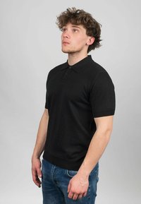 Svart kortärmad polo med vikbar krage, med treknappsslutning och ribbad nederkant, bärs tillsammans med blå denimjeans.