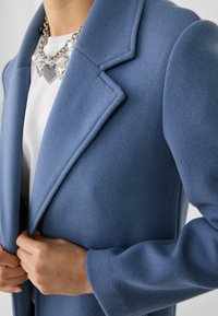 Modrý kabát z vlněné směsi s klopou s výřezem, strukturovanými rameny a měkkou texturou. Nošen přes bílé tričko a doplněn řetízkovým náhrdelníkem.