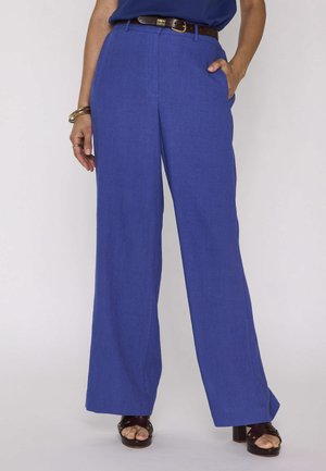 Donna che indossa pantaloni royal blue a gamba larga con cintura marrone, sandali scuri con tacco blocco aperto sulla punta e accessori al polso sinistro.