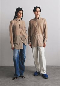 Due persone stanno fianco a fianco. Uno indossa una camicia oversize marrone chiaro e jeans blu, l'altro una camicia simile con pantaloni bianchi e scarpe blu.