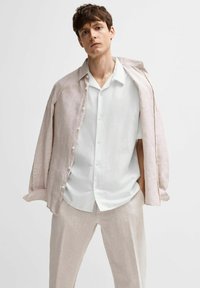 Camicia di lino rosa chiaro sopra una camicia bianca con bottoni, abbinata a pantaloni di lino beige chiaro. Texture morbide e vestibilità rilassata evidenti.