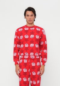 Felpa rossa con un motivo di rose rosa su tutta la superficie, colletto rotondo, maniche lunghe e vita aderente. Abbinata a pantaloni coordinati.