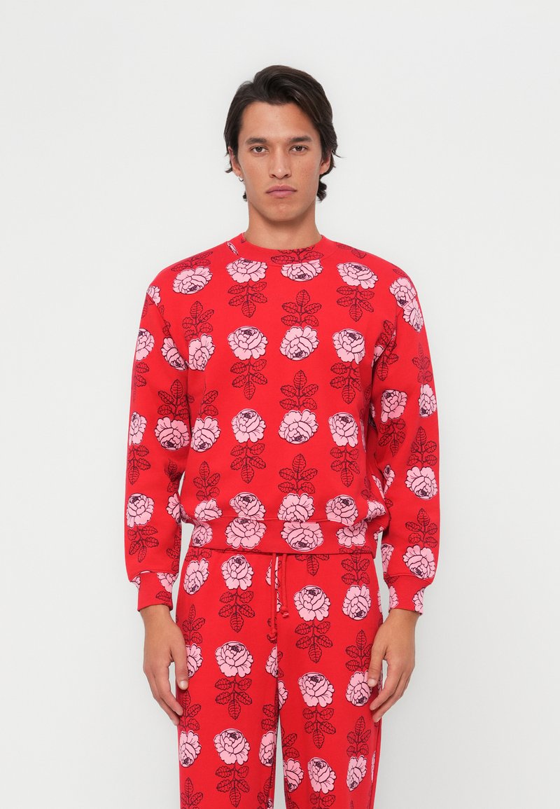 Felpa rossa con un motivo di rose rosa su tutta la superficie, colletto rotondo, maniche lunghe e vita aderente. Abbinata a pantaloni coordinati.