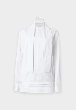Femme debout portant une blouse blanche à manches longues avec des liens verticaux et un pantalon noir à jambes larges avec des sandales noires à talons.