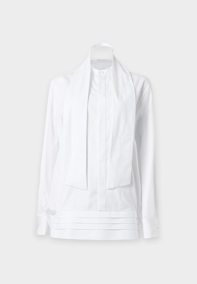 Blouse blanche à manches longues avec large cravate attachée et plis horizontaux près de l'ourlet, présentée sur un fond clair uni.