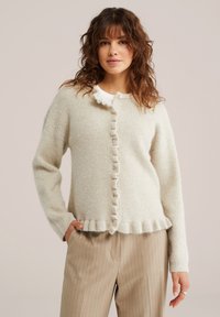 Cardigan bej deschis tricotat, cu margine cu volane, prevăzut cu închidere cu nasturi și un design texturat, cu dungi. Purtat peste o cămașă albă.