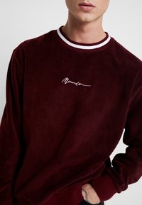 Burgunderrød fløjlssweatshirt med en hvidstribet rund hals, med broderet signaturdetalje på brystet.
