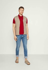 Colete acolchoado bege sobre uma camisa polo vermelha, combinado com jeans slim azul claro e sandálias castanhas. Design simples com um corte descontraído.