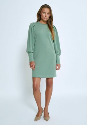 Mintgroene jurk met lange pofmouwen, ronde hals en een eenvoudig, aangesloten silhouet. Soepele textuur; gecombineerd met huidkleurige hakken.