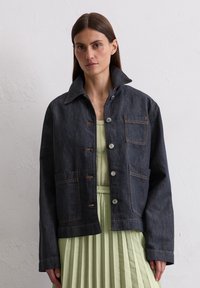 Dark denim jacket with a collar, five-button closure, front pockets, and orange stitching details, worn over a pleated light green dress.