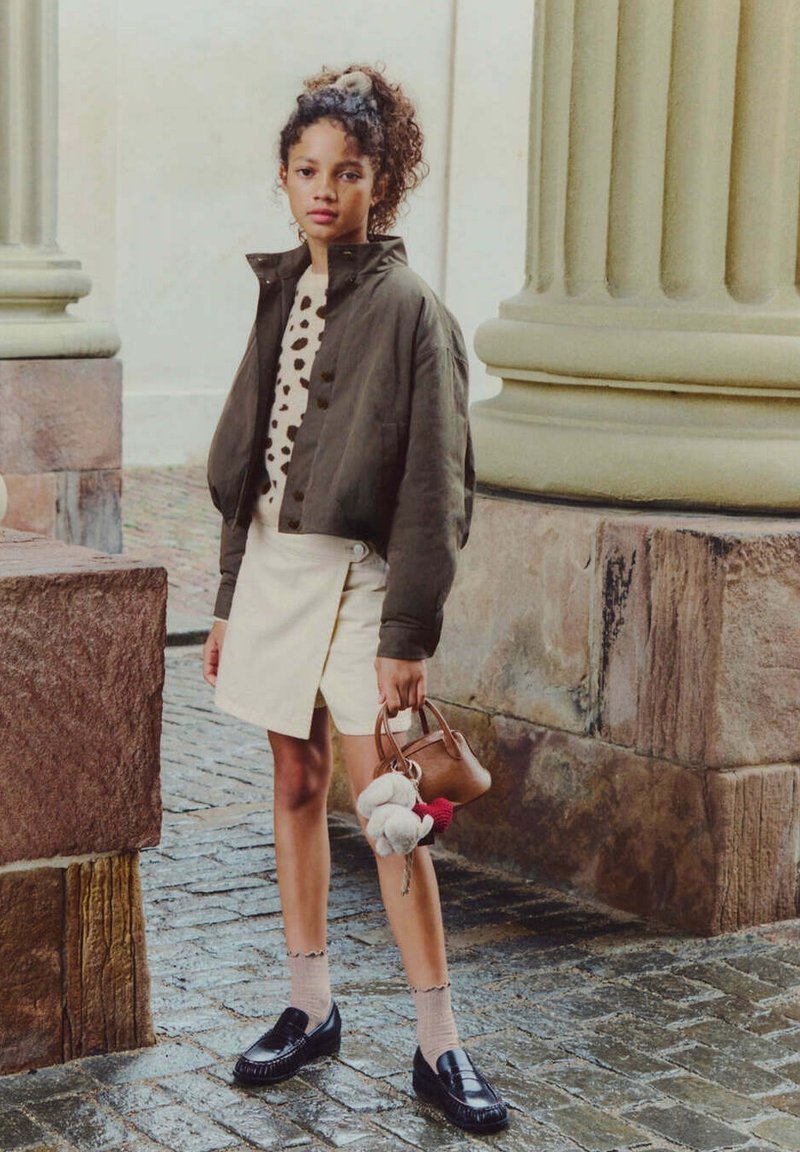 Jeune femme aux cheveux bouclés porte une veste sombre, un pull à pois, une jupe crème et des mocassins noirs, tenant un sac à main marron avec des porte-clés en peluche.