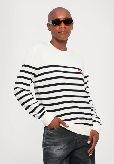 Person wearing dark sunglasses, white sweater with black horizontal stripes, and faded black jeans, standing with one hand in pocket.