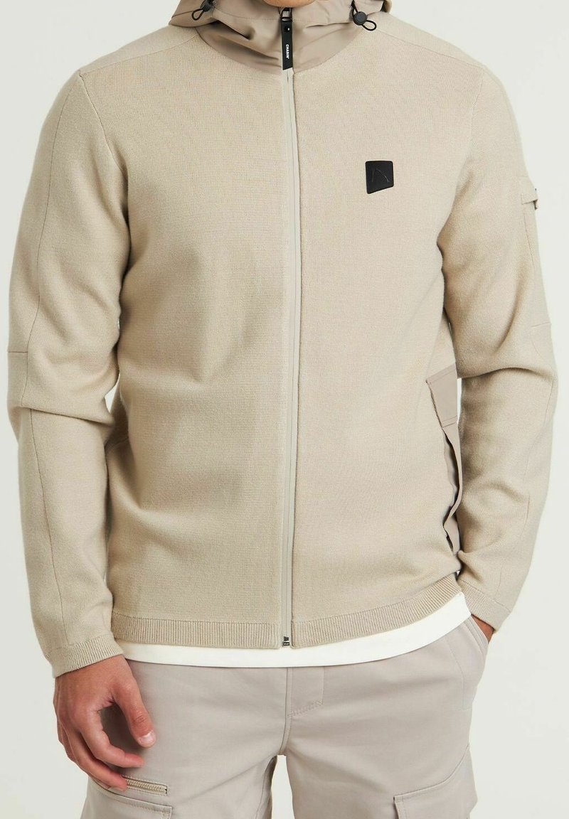 Homme portant un sweat à capuche beige zippé avec un logo noir sur la poitrine, pantalon beige et chemise blanche dépassant de l’ourlet, mains dans les poches.
