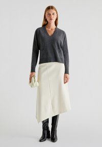 Donkergrijze V-hals trui met lange mouwen, gecombineerd met een asymmetrische, off-white rok. Accessoires zijn een lichtgekleurde clutch en zwarte laarzen tot de knie.
