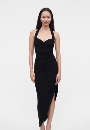 Femme debout et souriante, vêtue d'une robe noire sans manches à encolure américaine, décolleté en cœur et ourlet asymétrique, sur fond uni.