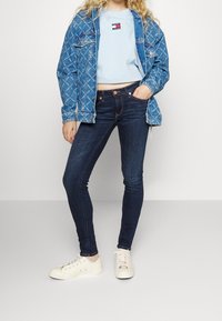 Chaqueta de mezclilla con patrón de diamantes, sudadera recortada de color azul claro con logo, jeans oscuros ajustados y zapatillas blancas rotas con un acento colorido.
