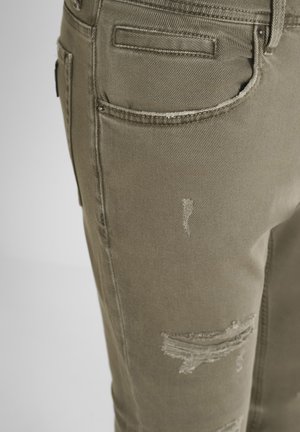 Nahaufnahme eines Paares khakifarbenen, gewaschenen Jeans, die die Details der Vorderpockets und die ausgefransten Stoffbereiche am Oberschenkel zeigen.