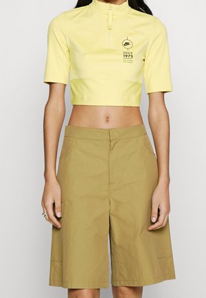 Woman wearing yellow cropped Nike zip-up top with "Since 1972" text and beige knee-length shorts, standing against a plain background.