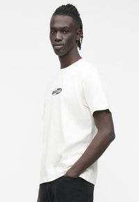 Valge puuvillane t-särk lühikeste varrukatega ja ümara kaelusega, mille rinnal on roheline ja musta värvi "Guess Jeans" logo.