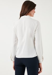 White long-sleeve blouse with a high collar, back zipper, and buttoned cuffs. Smooth texture with a tailored fit.
