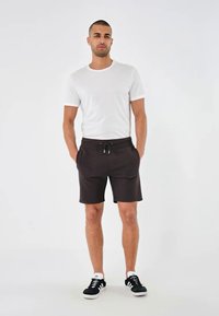 Hvit t-skjorte med kort erme og mørkebrune shorts med justerbar snøring, kombinert med svart og hvite joggesko. Ensfarget bakgrunn.