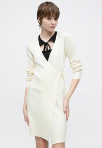 Strikket omslagskjole i off-white, med lange ermer, dyp V-hals, knyttet midje og en tettsittende silhuett. Teksturert ribbestrikket stoff, minimalistisk design.