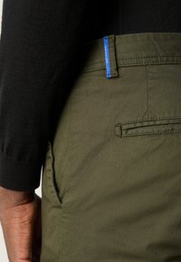 Olijfgroene broek met een gestructureerde afwerking, voorzien van een blauwe accent bij de tailleband en een zijzak. Slim fit ontwerp met een gepersonaliseerde afwerking.