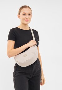 Borsa a tracolla beige chiaro in morbido tessuto, caratterizzata da una forma arrotondata e una tracolla regolabile, con un'etichetta del marchio sul frontale.
