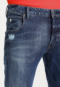 Tummansininen farkkuhousut, joissa on kuluneet yksityiskohdat, kuten repaleiset paikat ja hieno haalistuminen. Klassinen viisi-taskumalli metalliosilla.