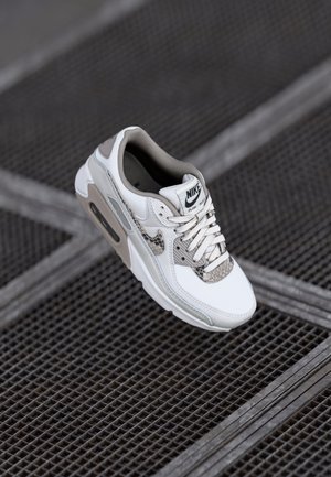 White and gray Nike Air Max sneaker with snakeskin pattern accents displayed on a dark metal grid floor.