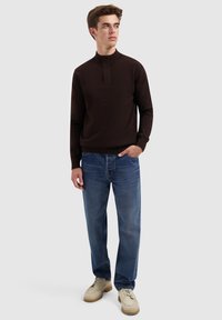 Junger Mann in dunkelbraunem Pullover, blauen Jeans und beigen Schuhen, der mit einer Hand in der Tasche vor einem schlichten hellen Hintergrund steht.