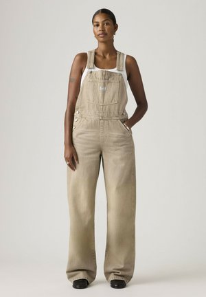 Donna con capelli scuri raccolti in uno chignon, che indossa una salopette beige a gamba larga e una canottiera bianca, in piedi con una mano in tasca davanti a uno sfondo semplice.