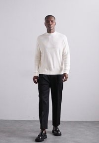 Pulover tricotat crem, pantaloni negri eleganți și pantofi negri lucioși; linii curate și un design minimalist pe un fundal neutru.