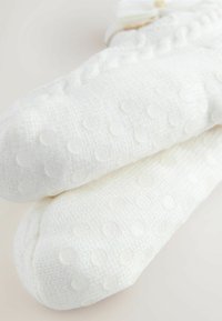 Bijele teksturirane čarape s mekanim pletenim uzorkom s podignutim točkicama i glatkom završnom obradom. Uključuju dekorativni ukras u obliku mašne.