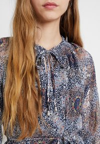 Jonge vrouw met lang lichtbruin haar, gekleed in een doorschijnende blouse met bloemenpatroon en een gestrikte halslijn met zilverkleurige uiteinden.